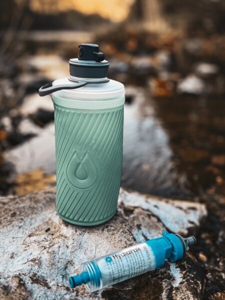 Skládací láhev Flux™ HydraPak® s nasazeným filtrem na vodu HydraPak® (28 mm) položená na kameni u potoka. Foto: Rigad