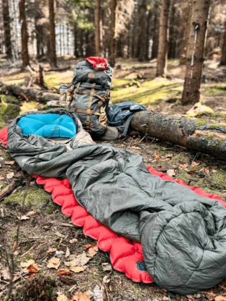 Zelenošedý spacák na červené nafukovací karimatce s modrým polštářkem a batohem v lese.