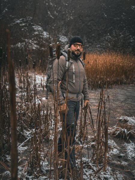 Muž v zimním outdoorovém oblečení s batohem v zasněženém mokřadu.