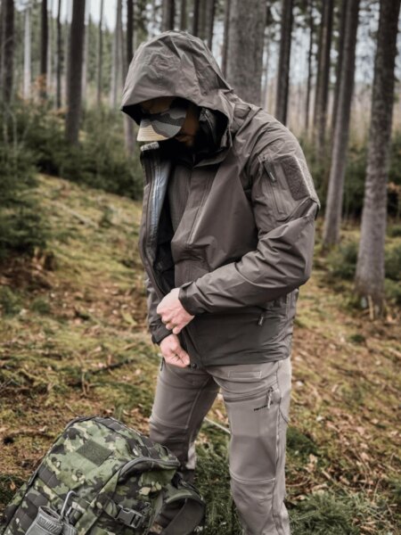 Muž v nepromokavé bundě Helikon-Tex Chinook s kapucí vedle maskovacího batohu v lese.