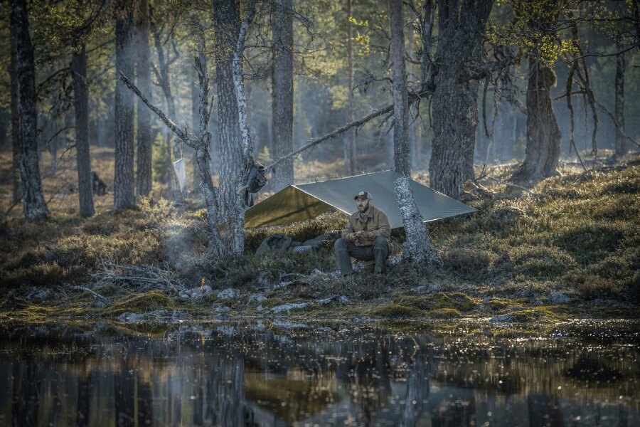 Nízkoprofilový tábor — tarp v zemitých barvách splyne s okolím, oheň malý a krytý. Odchod = LNT: místo srovnat, žádné stopy. Foto: Helikon-Tex, Rigad
