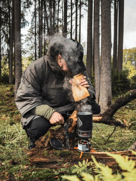 Muž v zateplené bundě klečí v lese u vařiče Jetboil, z něhož stoupá pára. Připravuje teplé jídlo nebo nápoj jako zdroj energie a tepla během podzimní výpravy. Foto: Rigad