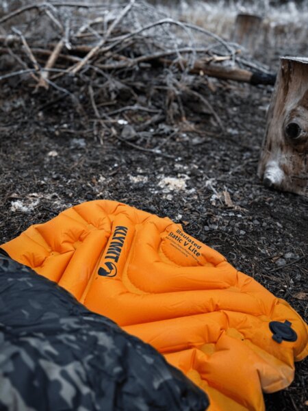 Nafukovací karimatka Klymit Insulated Static V Lite v oranžové barvě na zemi u ohniště, částečně zakrytá spacákem s maskovacím vzorem. Izolace od chladu a vlhkosti při spaní v terénu. Foto: Rigad