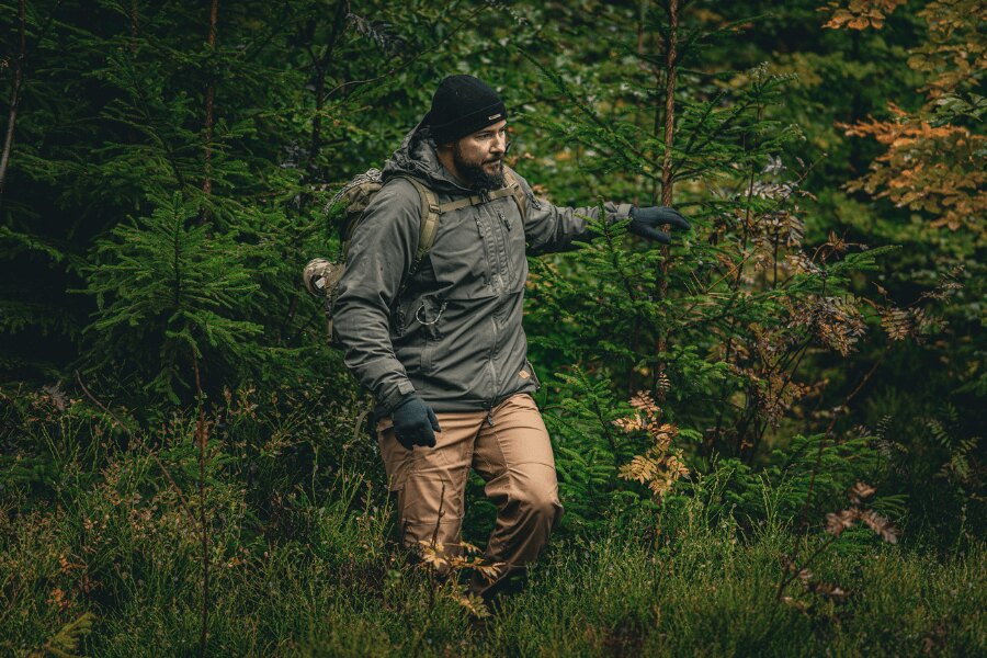 Muž v podzimním lese v šedé zimní bundě Yukon s izolací Climashield™ Apex® od Helikon-Tex®, čepici a rukavicích, s batohem; ukázka správného vrstvení a ochranné svrchní vrstvy do vlhka a větru. Foto: Rigad