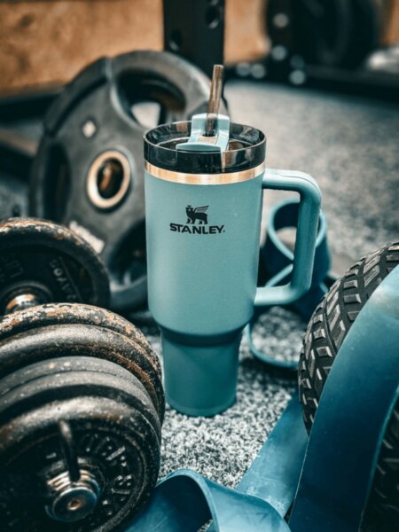 Termohrnek Stanley s brčkem položený mezi činkami a fitness vybavením.